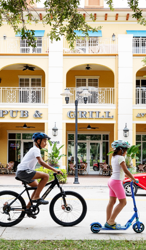 Children riding a bike and scooter past the Ave Maria town center pub and grill